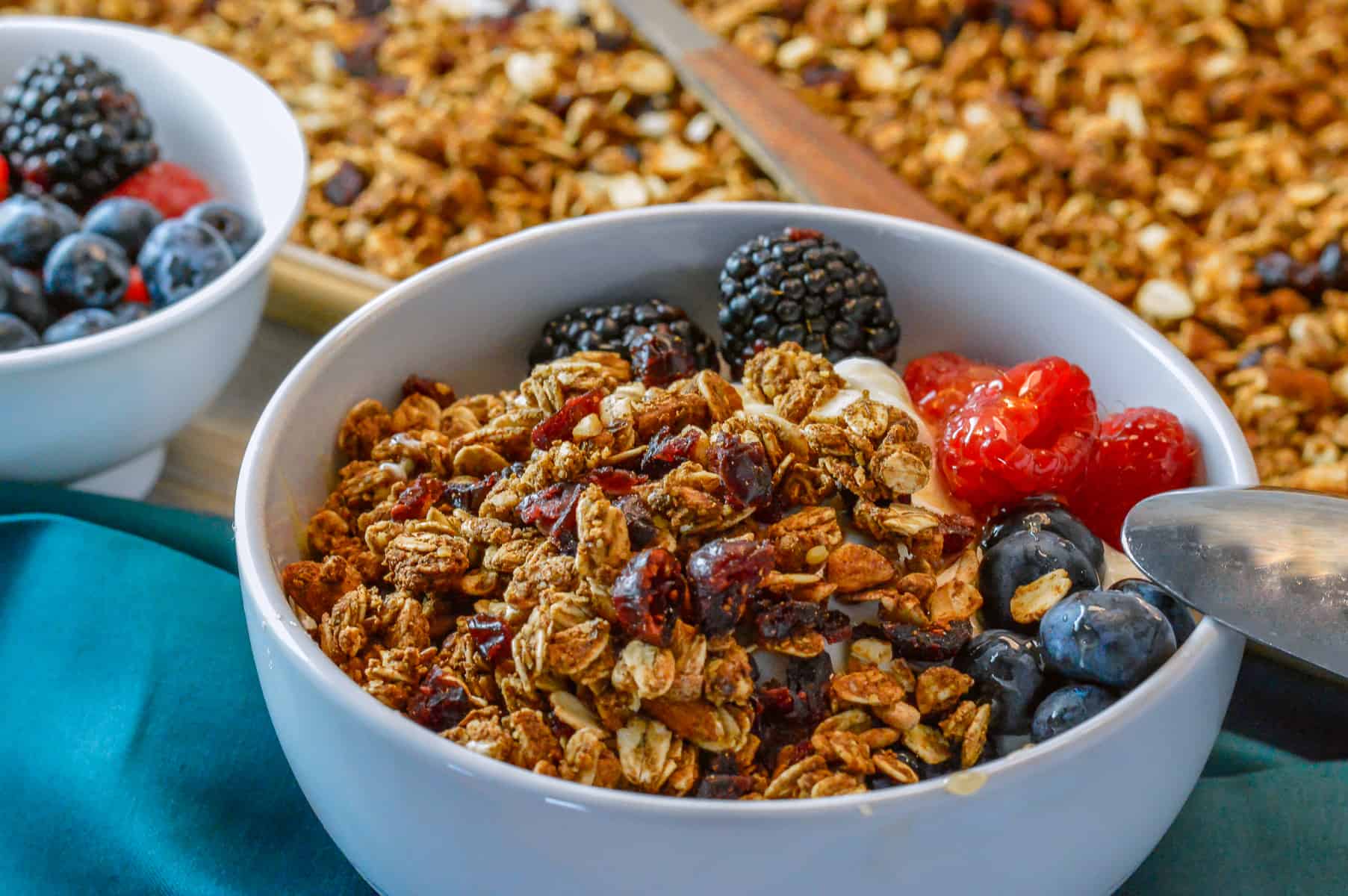 bowl of yogurt with granola and berries with more berries to the left and sheet pan of baked granola in background