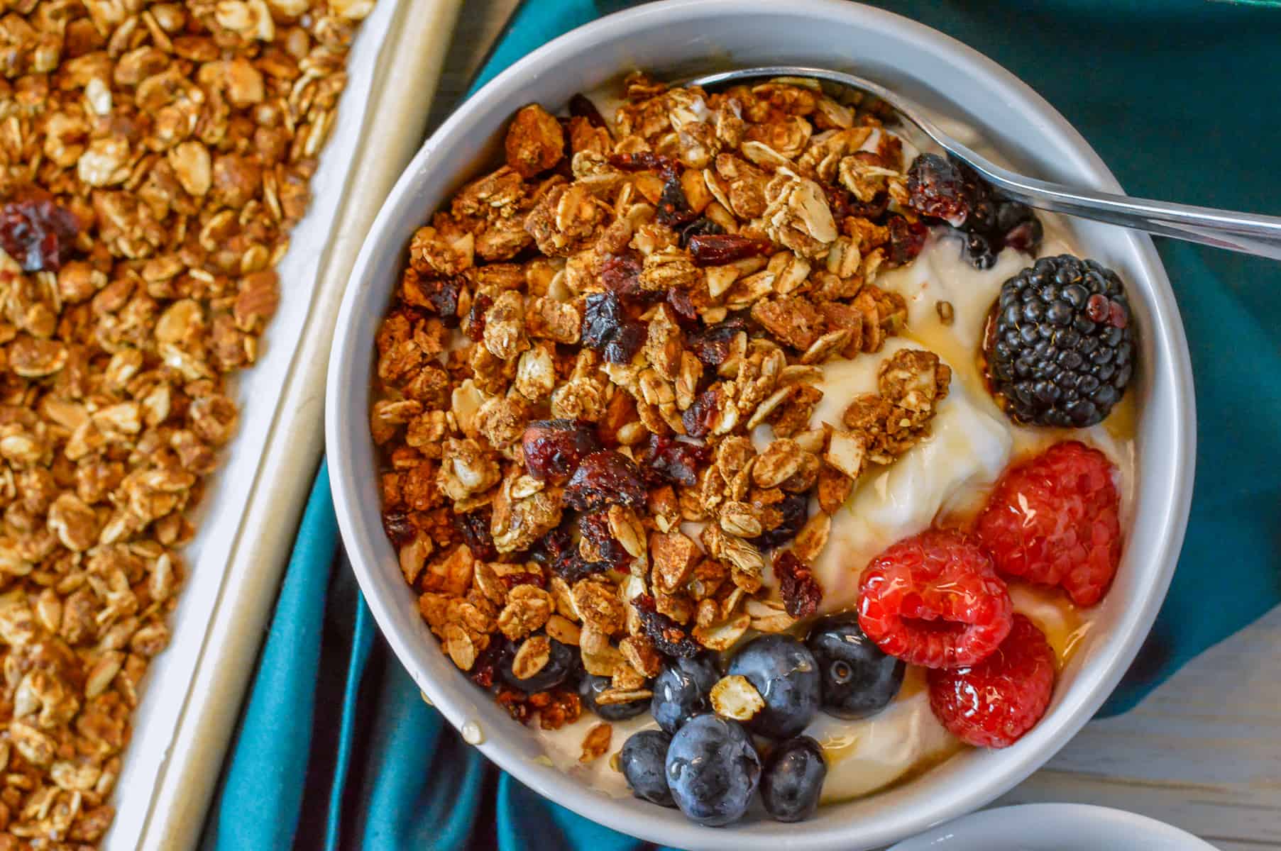 Over-top view of white bowl with yogurt, honey, berries and homemade granola on teal napkin with sheet pan of granola on the side.