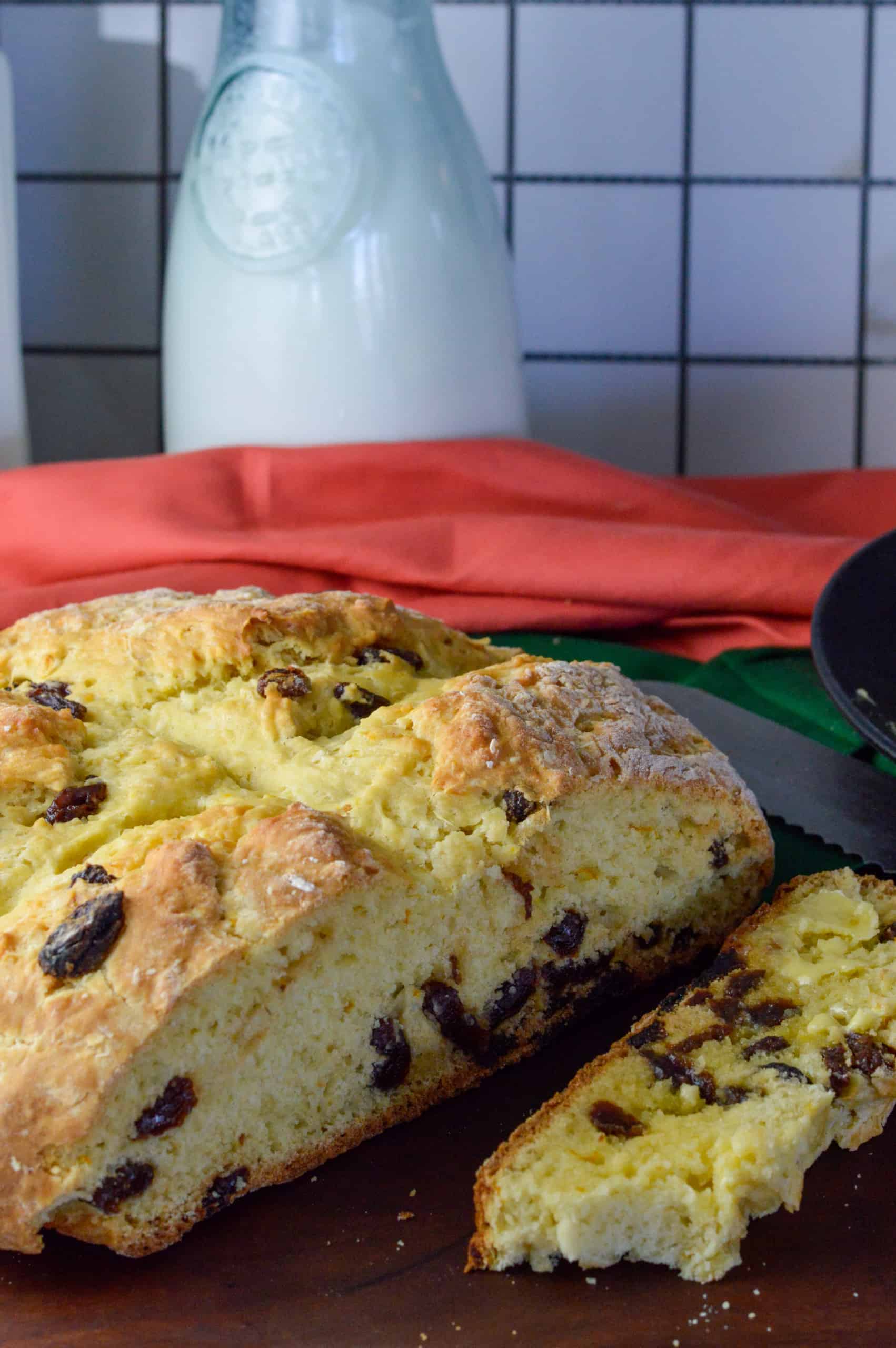 Bread loaf with raisins with slice cut off and buttered, orange and green napkins and buttermilk in background