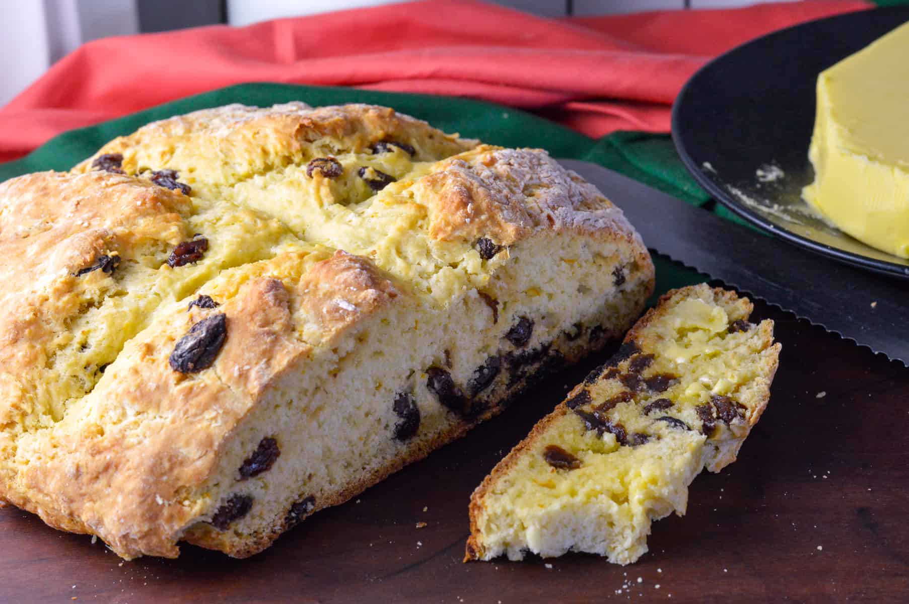 Irish Soda Bread with Raisins, sliced off side and slice buttered with bread knife, butter and green and orange napkins and buttermilk in background