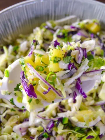 Up close shot of pineapple slaw with jalapeno, red onion and cilantro on white spoon spatula with bowl of slaw in background on wooden counter