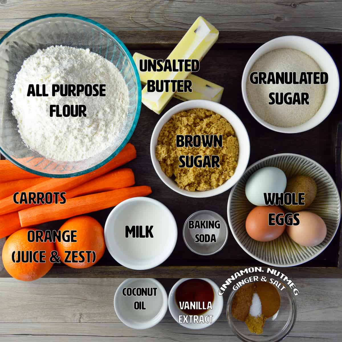 Ingredients for Orange Carrot Cake shown on wooden tray on counter: flour, butter, sugar, brown sugar, carrots, oranges, milk, baking soda, coconut oil, vanilla extract, eggs and spices