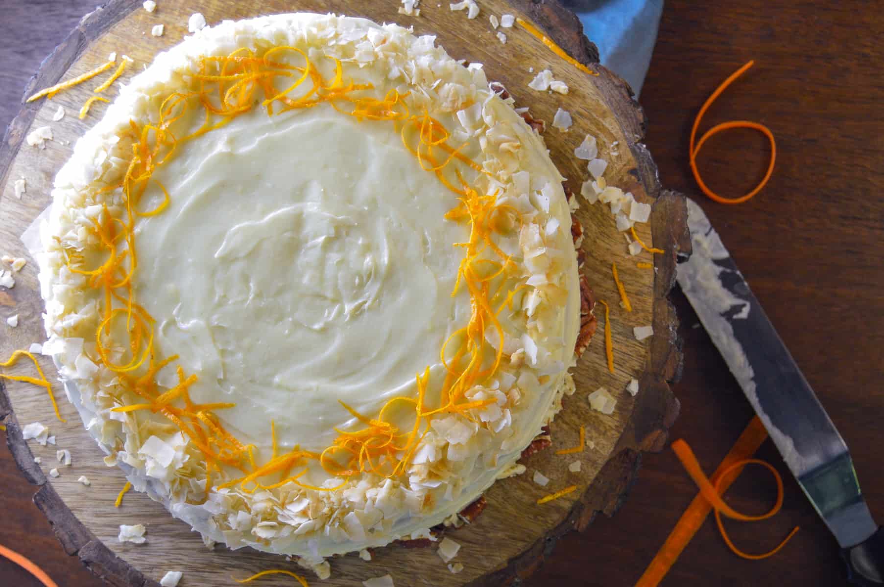Over top view of orange carrot cake on wooden cake board with coconut flakes, pecans and orange zest. carrot strips in background with frosting spatula