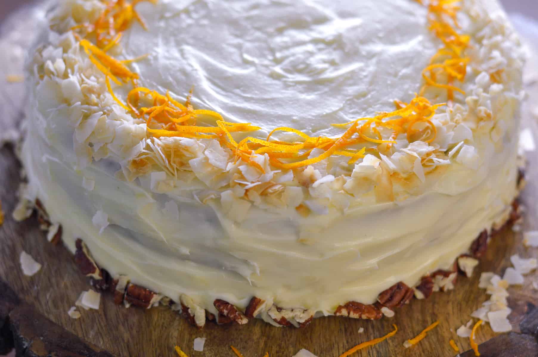 Close up of frosted carrot cake with roasted pecans on bottom edge, coconut flakes on top edge with orange zest all on wooden board