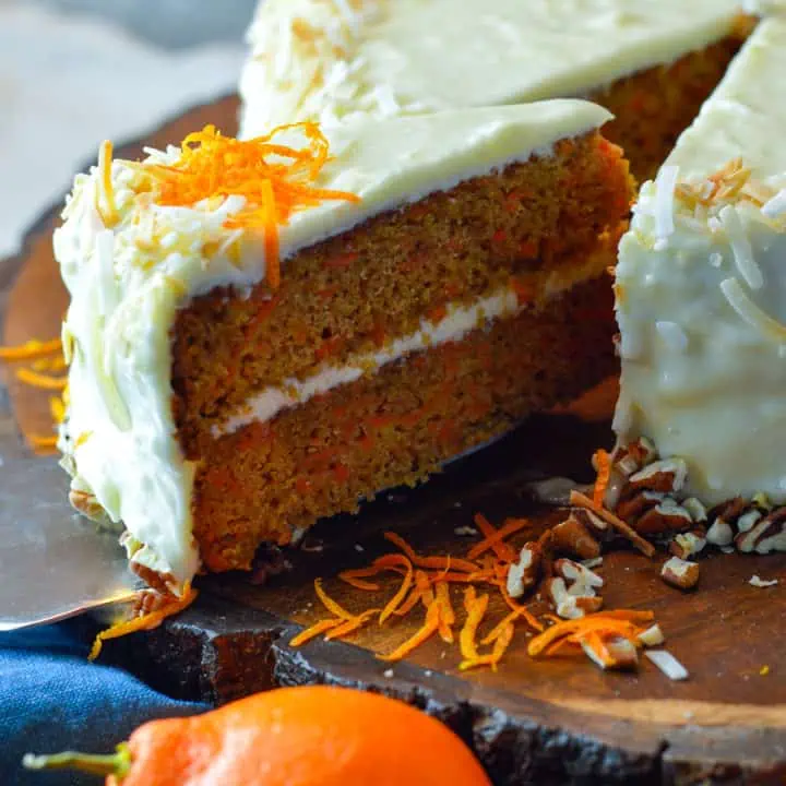 Orange Carrot Cake slice being grabbed with a cake spatula from the cake with orange in front and zest to the side.