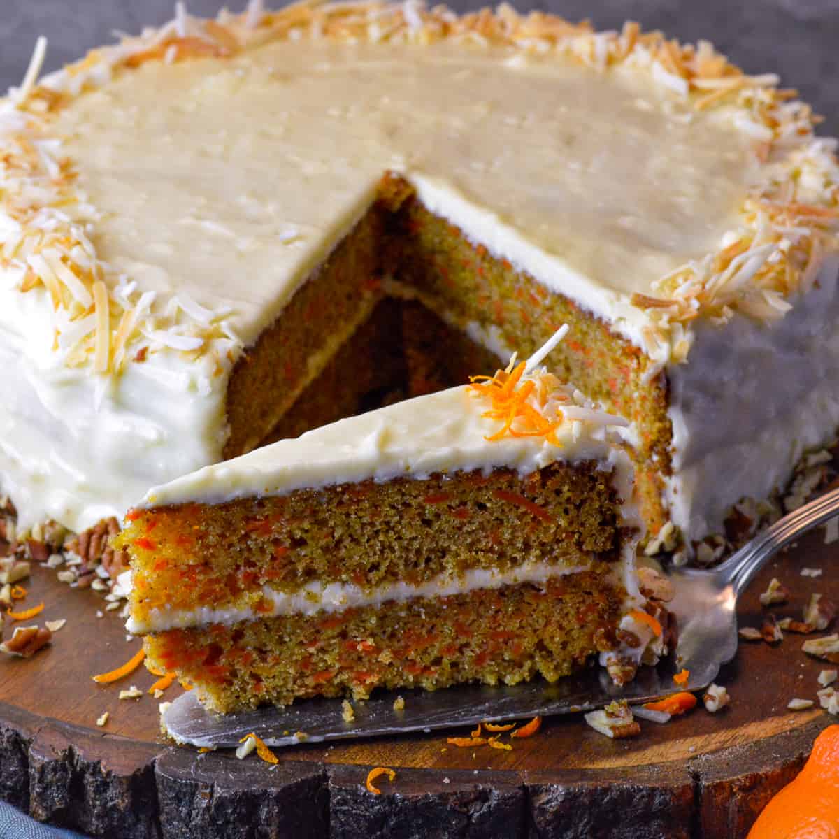 Orange Carrot Cake slice on silver cake spatula with roasted pecans, coconut flakes and orange zest with cream cheese frosting on wooden board