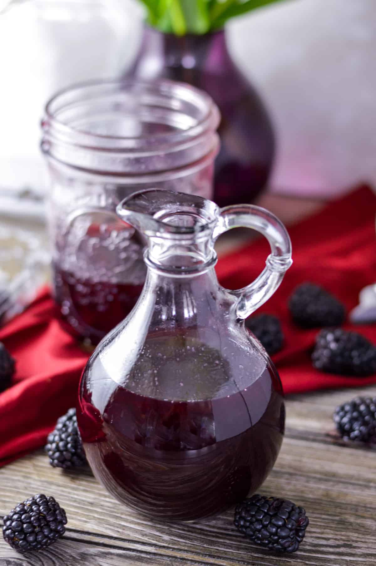 Blackberry Simple simple syrup in glass syrup holder with handle, mason jar, berry container and flower vase in background, blackberries around on counter