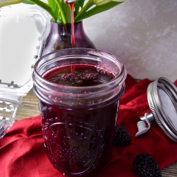 Close up drip of blackberry simple syrup in mason jar with berry container to side and pouring lid to the right on red napkin