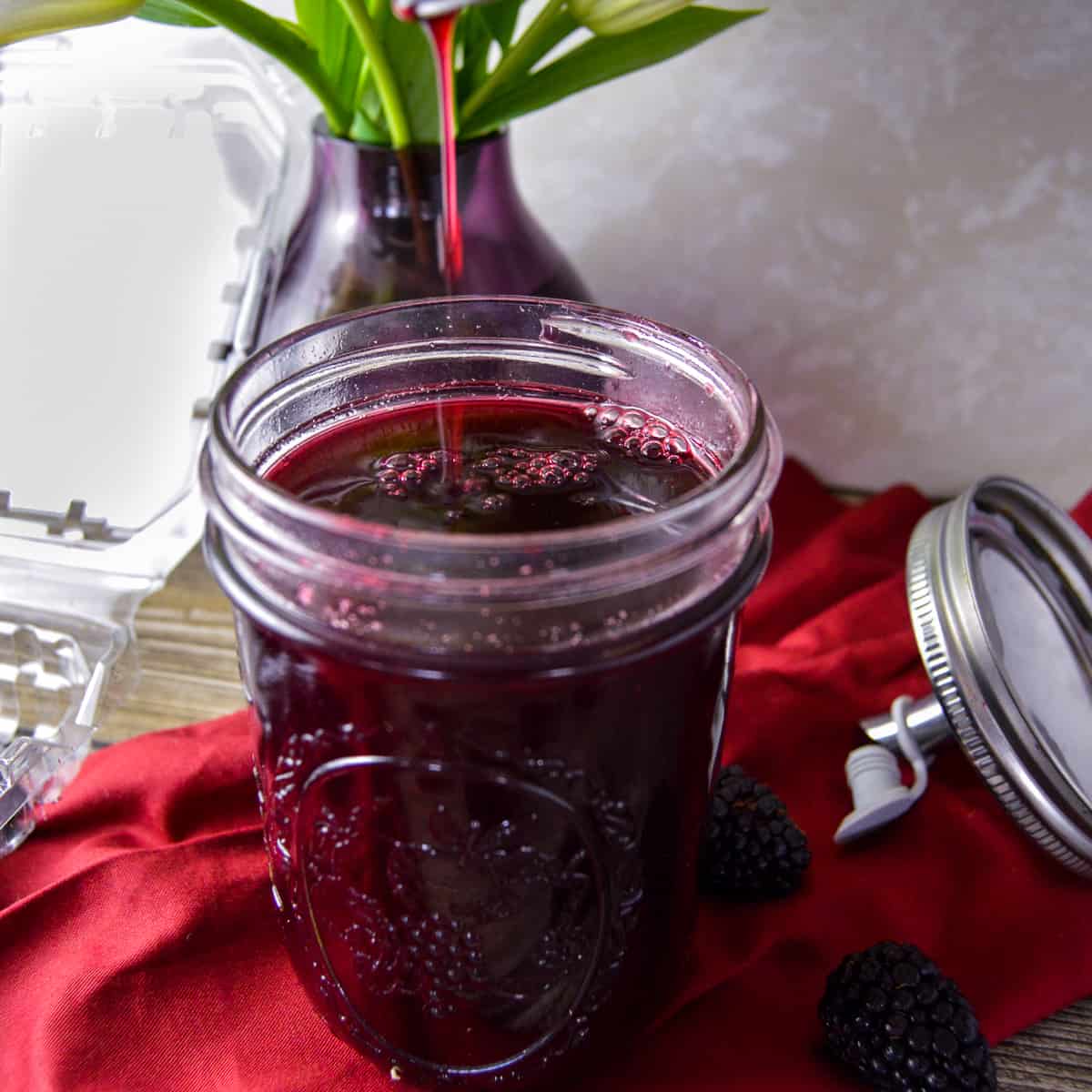 Blackberry simple syrup in mason jar being dripped from above jar that's over a red napkin 