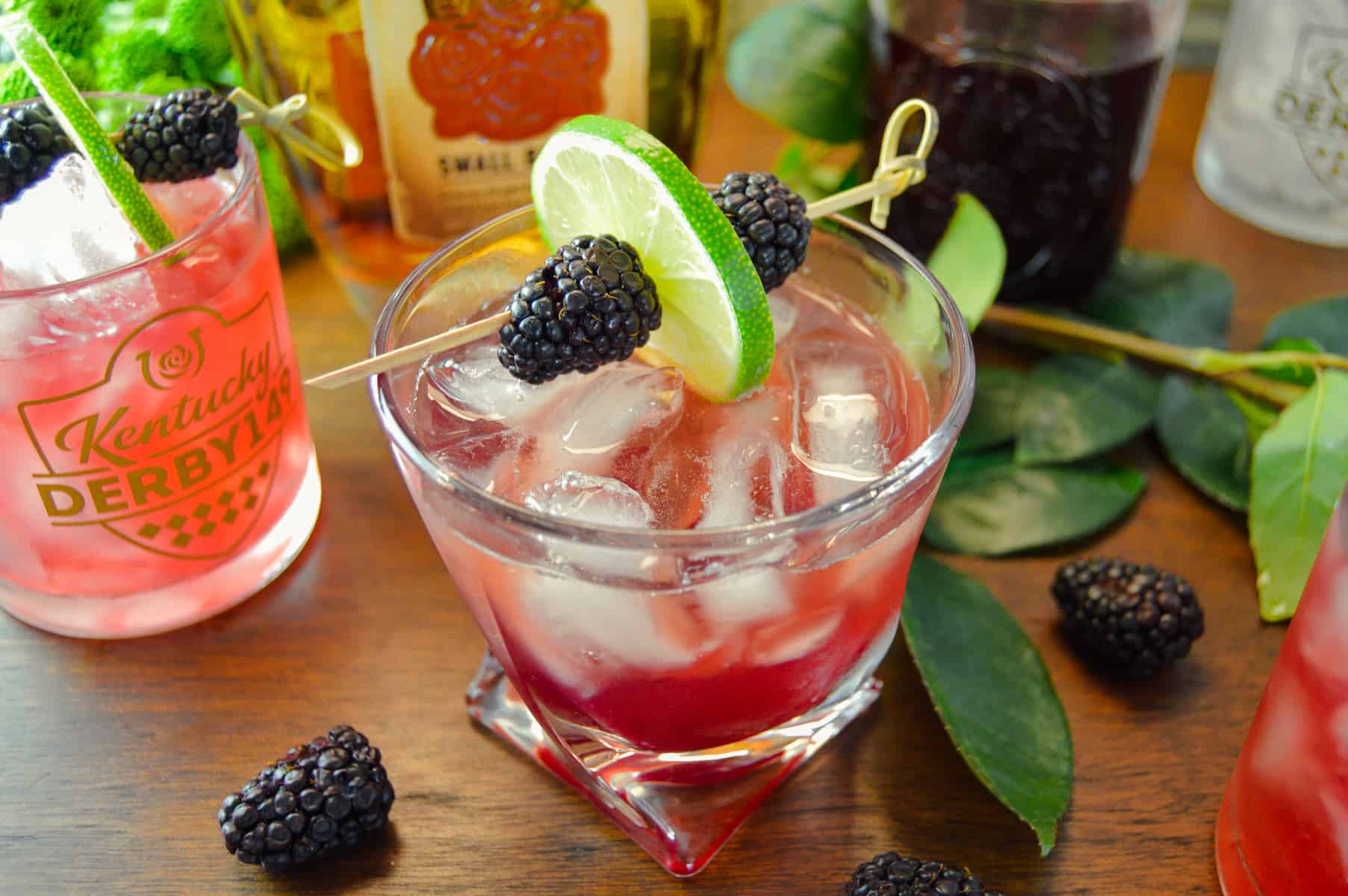 KY bourbon cocktails with blackberries around and pink rose, garnished with lime and blackberries on cocktail pick