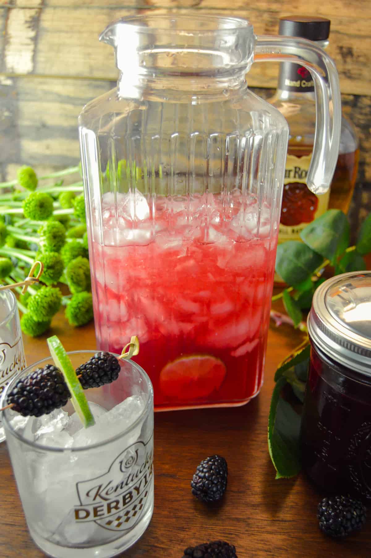 Glass Pitcher of Kentucky Blackberry Bourbon cocktail for a party with ice filled glasses with garnish and blackberry syrup in mason jar to the right 