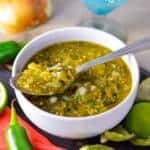 Spicy Salsa Verde in white bowl with a spoon of the salsa on top resting on dark wooden board by red nakin and tomatillos and jalapeno, onion and margarita glas