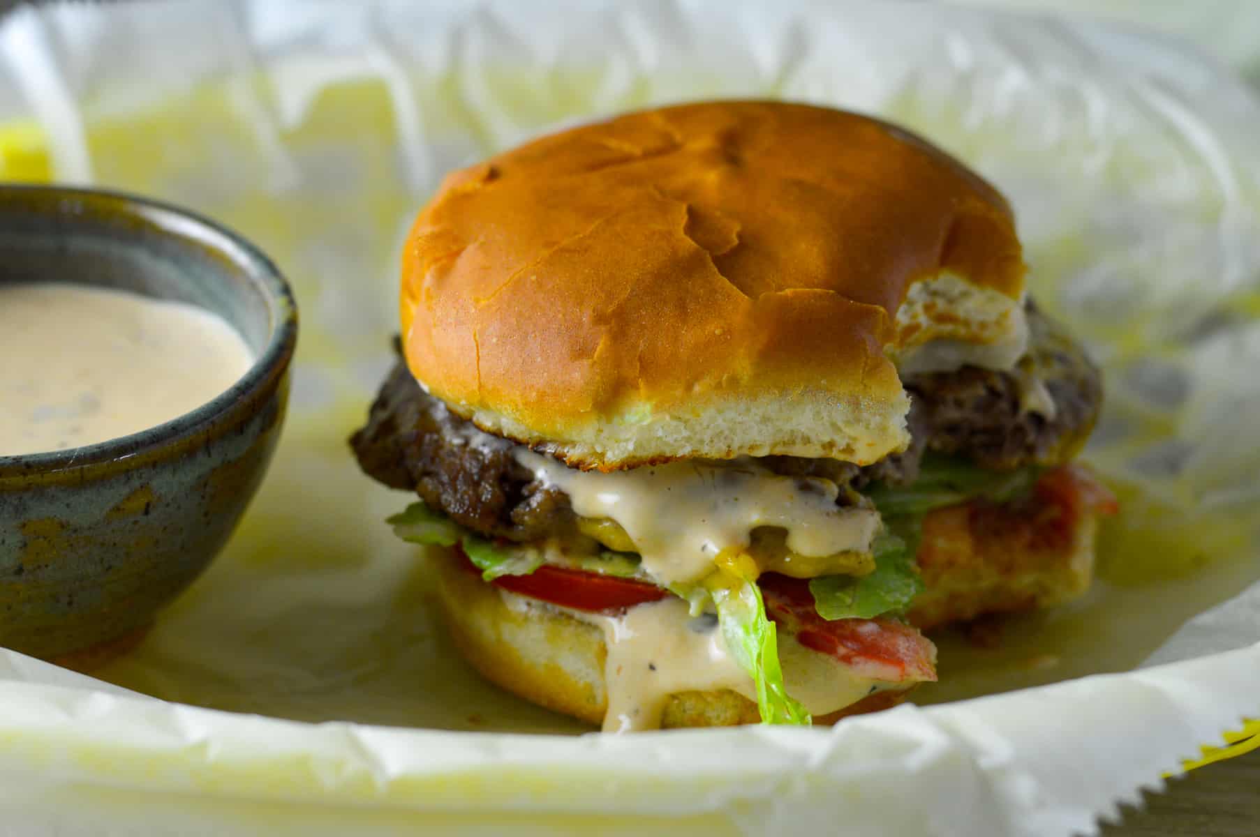 In-n-out-style smashed burger with bites taken out of it on a wax paper lined yellow burger basket with a small ceramic bowl of burger sauce on the side