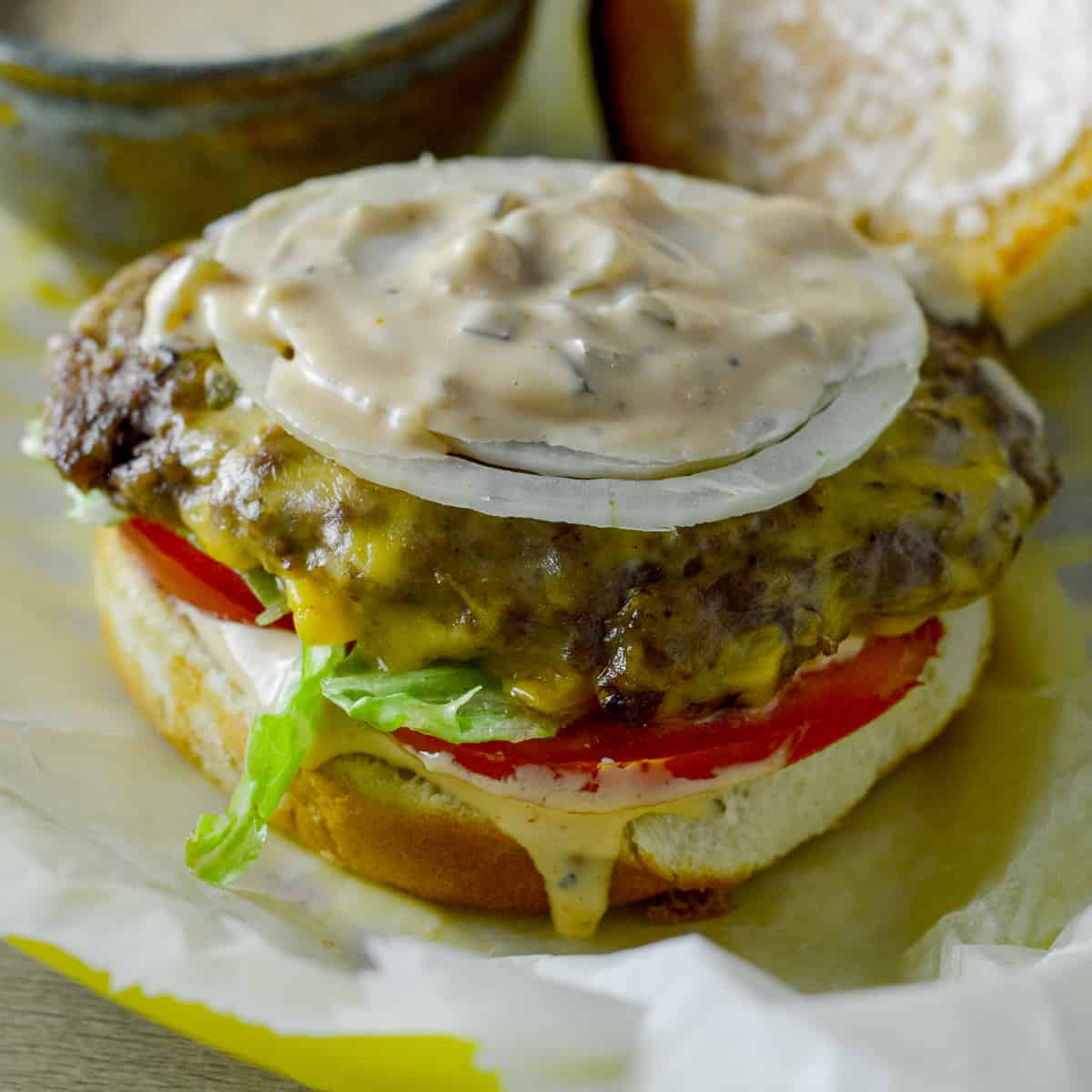 Open burger on wax paper over basket with sauce, tomato, lettuce, beef patty and onion rings, topped with in-n-out style homemade burger spread and top of bun on side and a small ceramic bowl with more sauce on the side