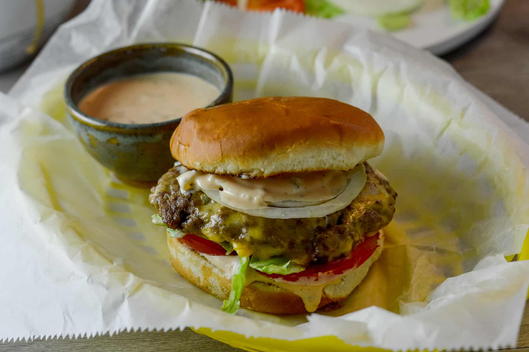 Full burger with all the toppings and toasted bun with extra sauce on the side on a wax paper covered yellow burger basket 