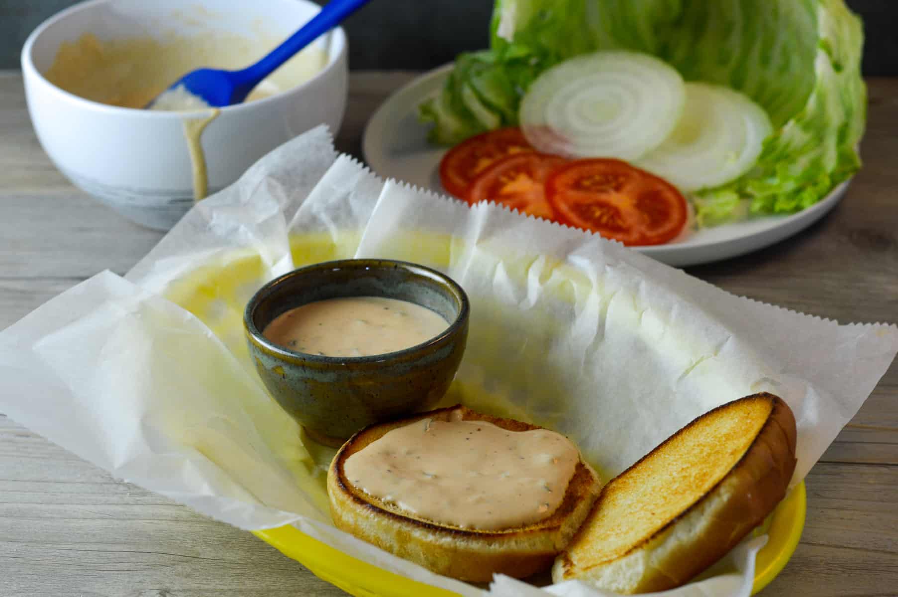 homemade burger sauce spread in a small bowl in basket with a toasted bun beside with burger sauce spread on the bottom of an open bun, more sauce in white bowl in background with some lettuce, tomato and onion in the background
