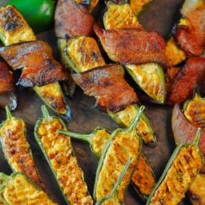 Smoked Jalapeno Poppers, bacon wrapped on top and plain below them on a wooden board with a raw green jalapeno in the upper left.