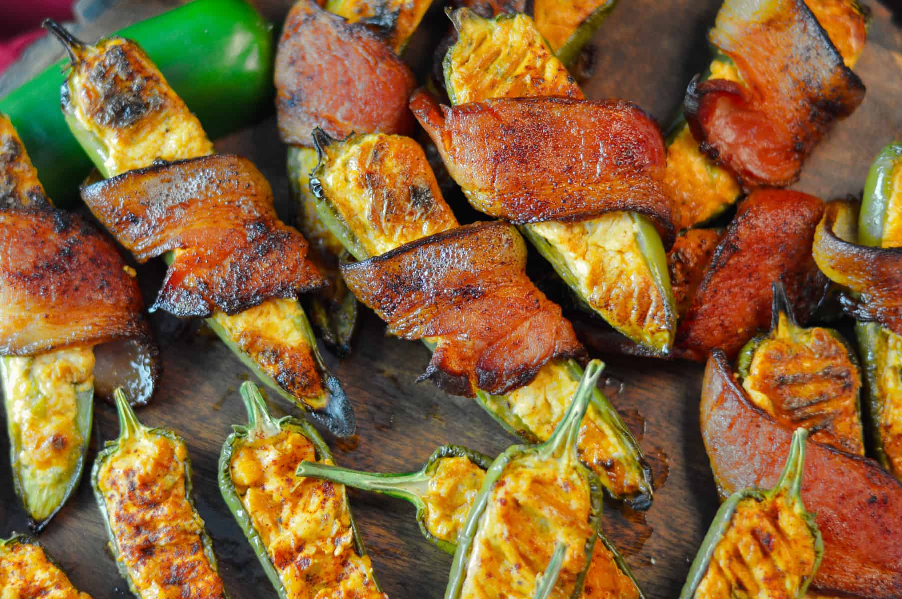 Close of up bacon-wrapper and some plain jalapeno poppers on a wood board from the top with a raw jalapeno in the top left side. 