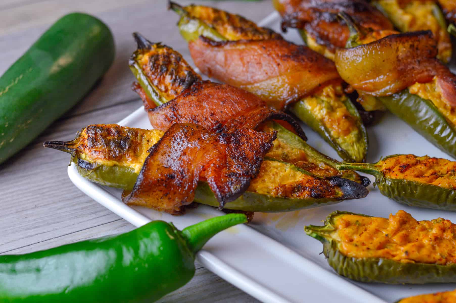 Bacon-wrapped smoked jalapeno poppers on a white platter with some raw jalapenos to the left side.