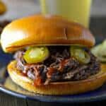 Easy Instant Pot Shredded Beef on a buns with a drizzle of bbq sauce and pickled jalapenos on a blue plate, pickles to the side and a beer and another beef sandwich behind