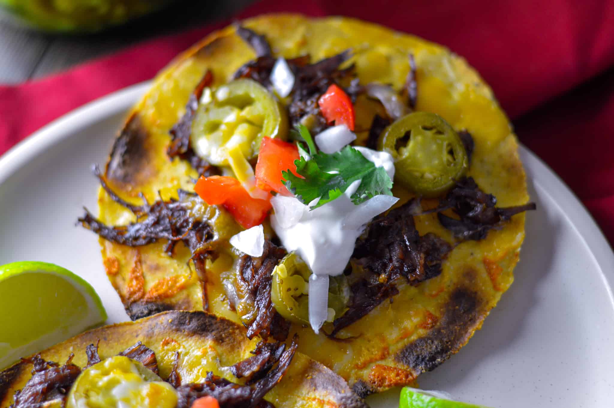 Shredded Beef on Tostada with a dollop of sour cream, pickled jalapeno, and tomato on white plate with lime wedge and red napkin beneath