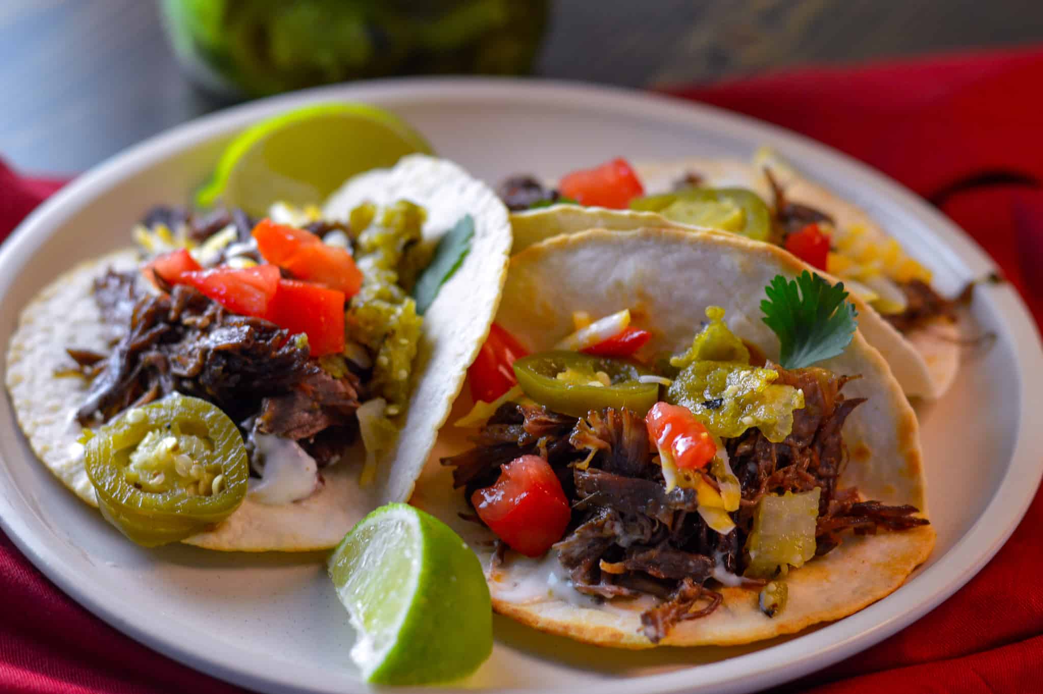 3 beef tacos with some cheese, jalapeno, tomato and cilantro on white plate over red napkin and pickled jalapenos in the background