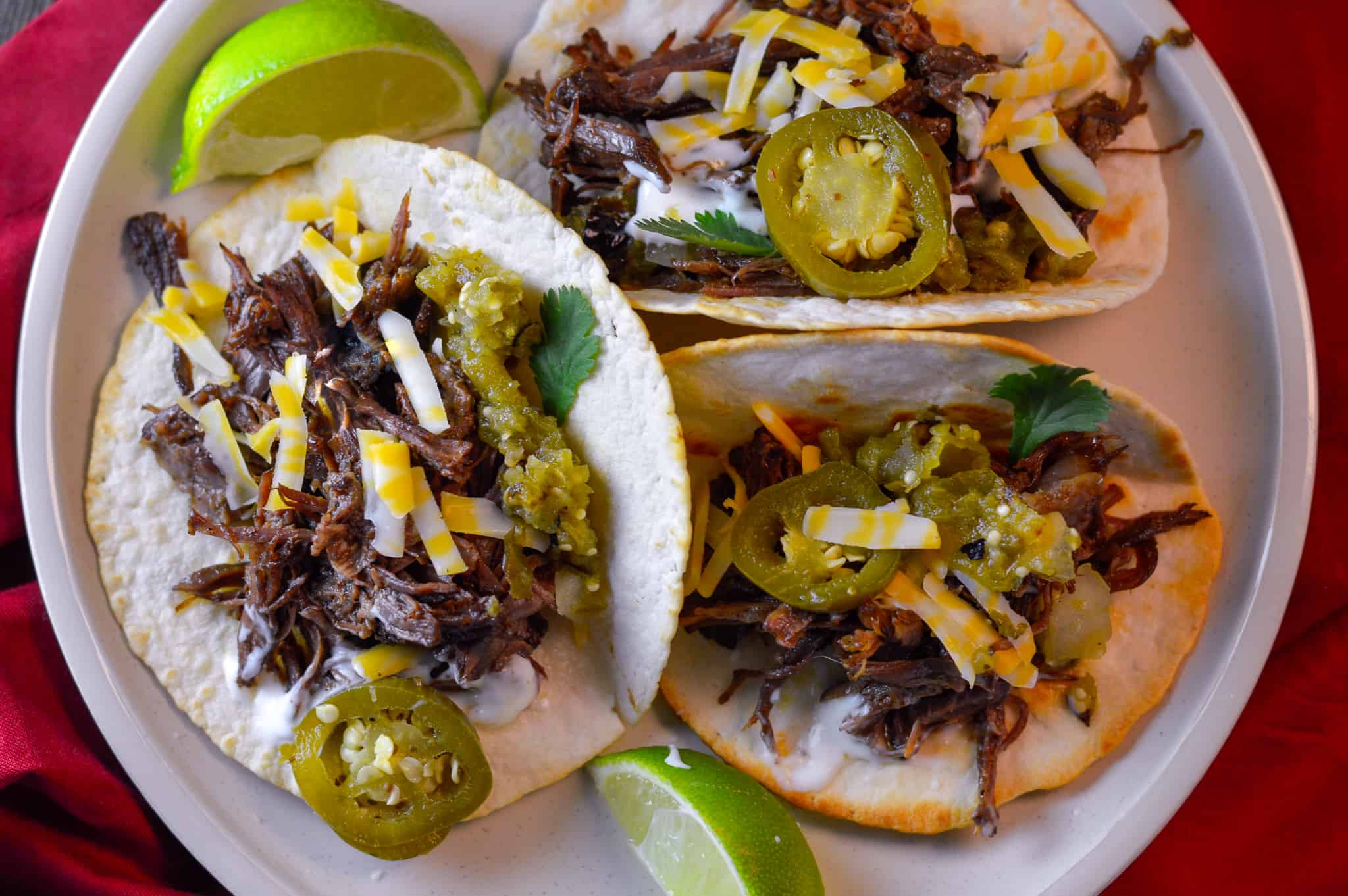 3 Shredded Beef street Tacos with some shredded cheese, salsa verde and pickled jalapeno on white plate with lime wedges