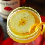 Up close shot of Orange Apple Cider Cocktail in coupe glass over red napkin with bourbon bottle, cider and orange seltzer in the background