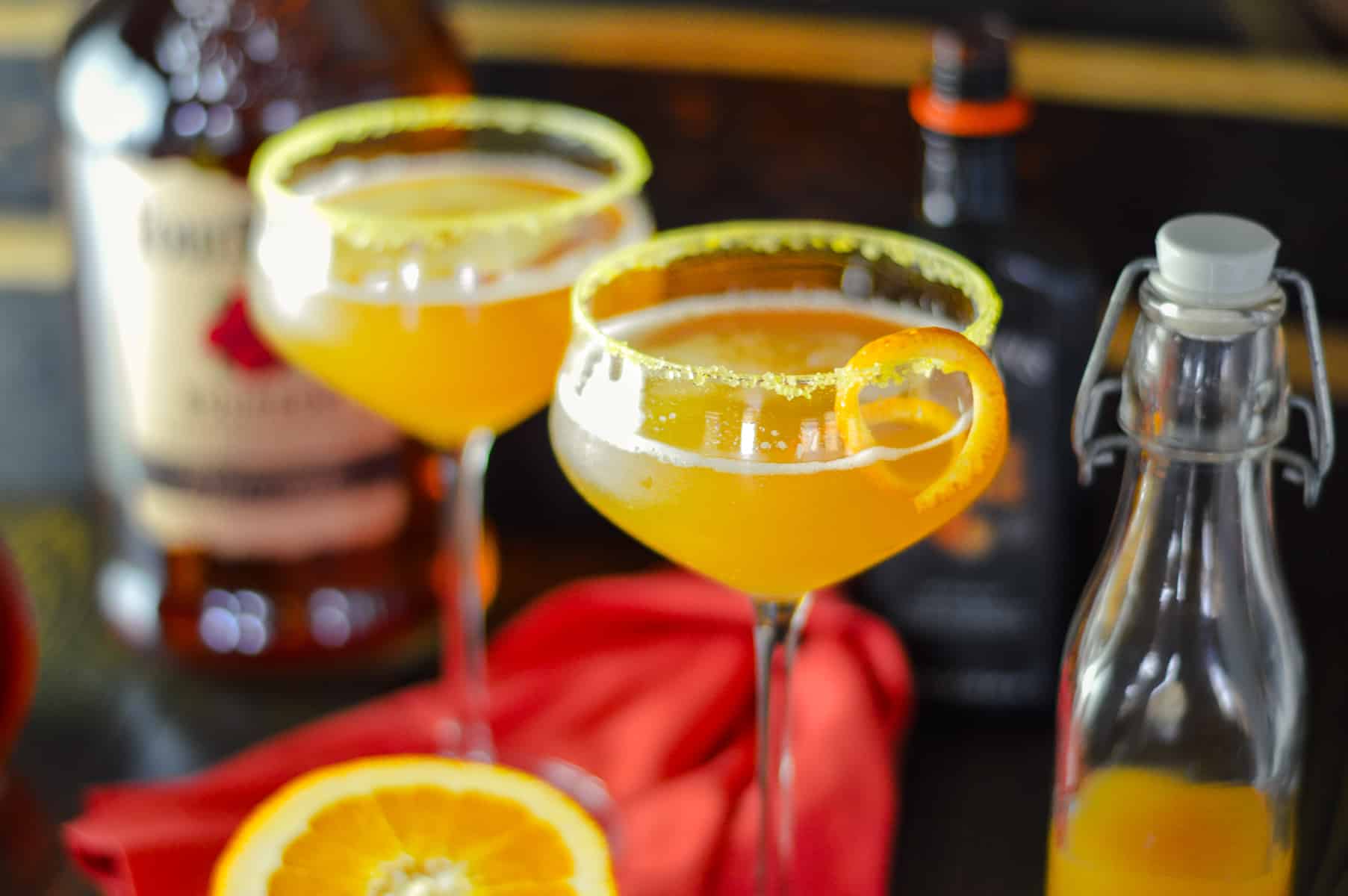 2 coupe glasses with orange apple cider cocktail on red napkin with half an orange in front, cider to the right, orange bitters behind and bourbon bottle to the left