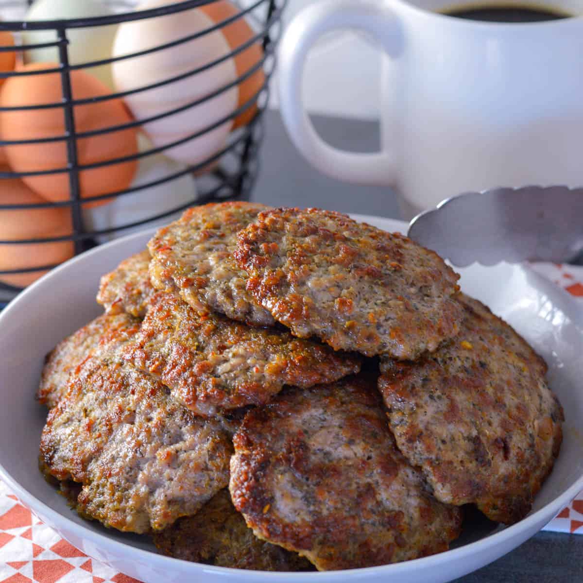 Stack of country breakfast sausage patties in a white serving bowl in red and white napkin with silver tongs, white coffee mug and basket of fresh eggs in background
