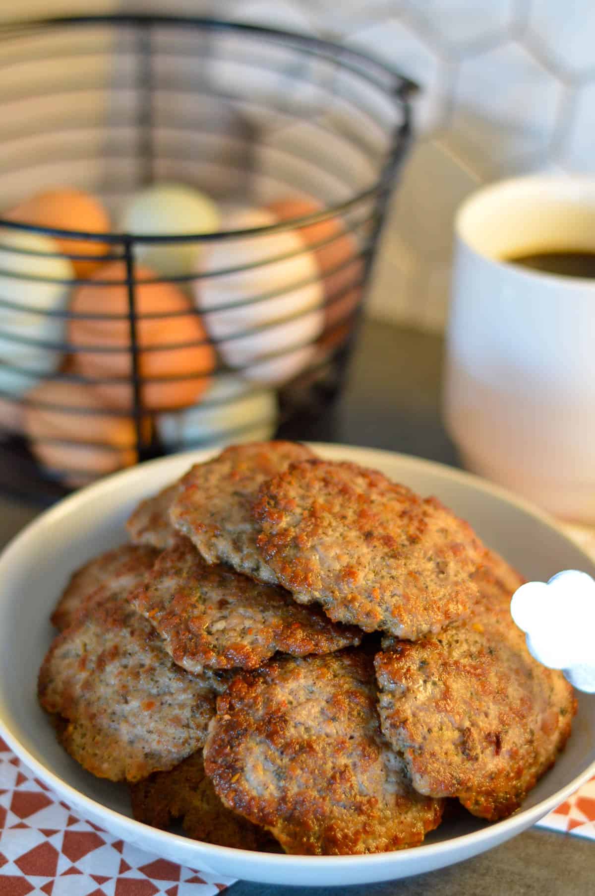 Breakfast Sausage Patties stacked in a white serving bowl over red and white napkin with basket of eggs and coffee cup in background