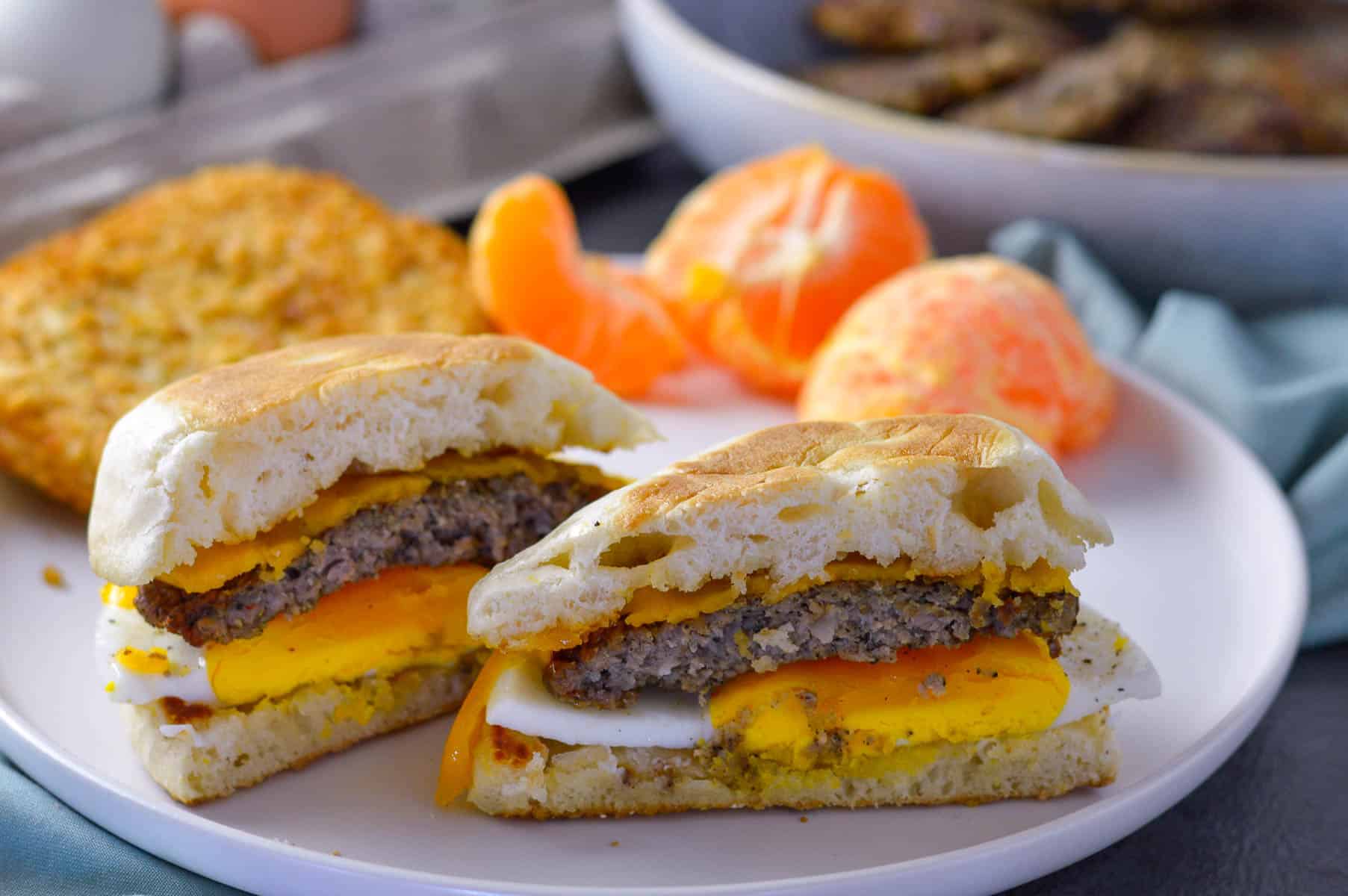 Breakfast sausage sandwich with fried egg and cheese on English muffin cut in half with hash brown patty and mandarin on white plate. Carton of fresh eggs and bowl of sausage patties in background