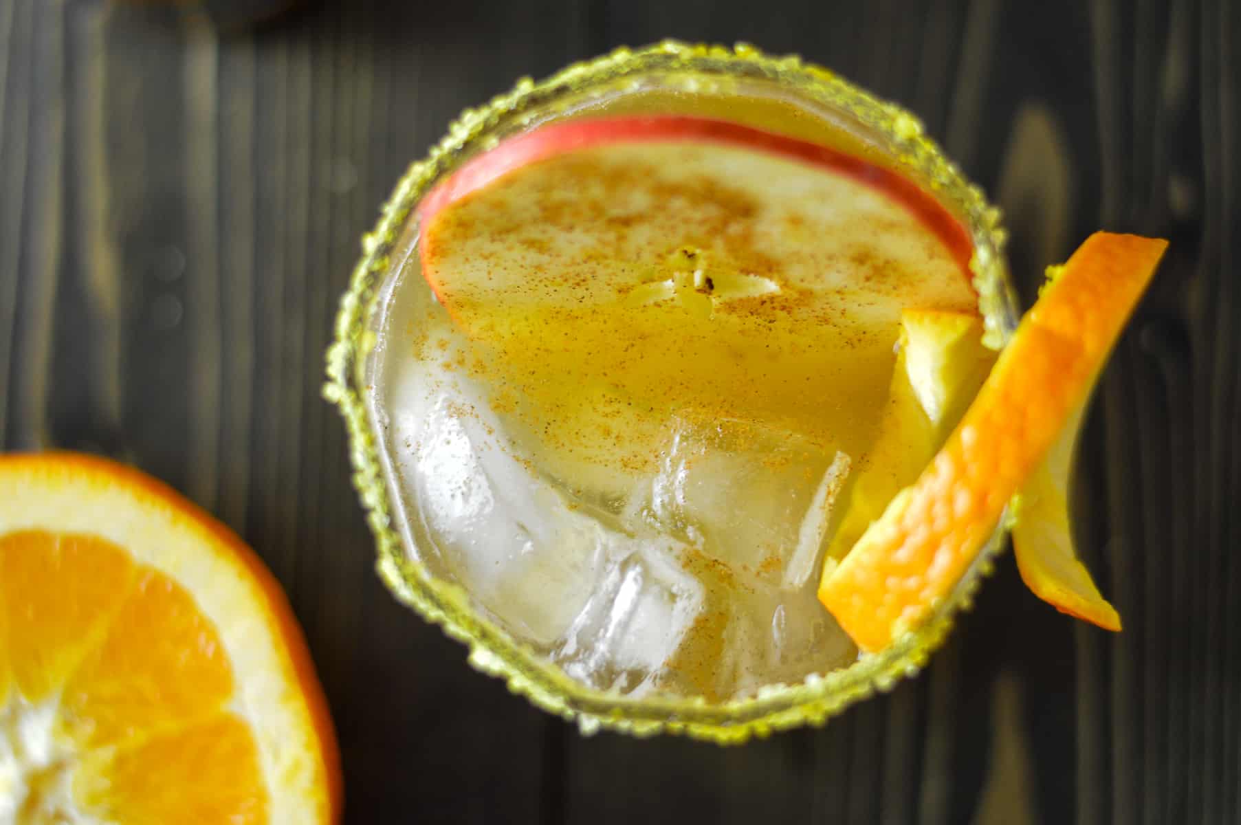 Over top view of apple cider cocktail on rocks with thin apple slice with cinnamon, orange peel twirl and some allspice in cocktail mixture with a half orange to the left