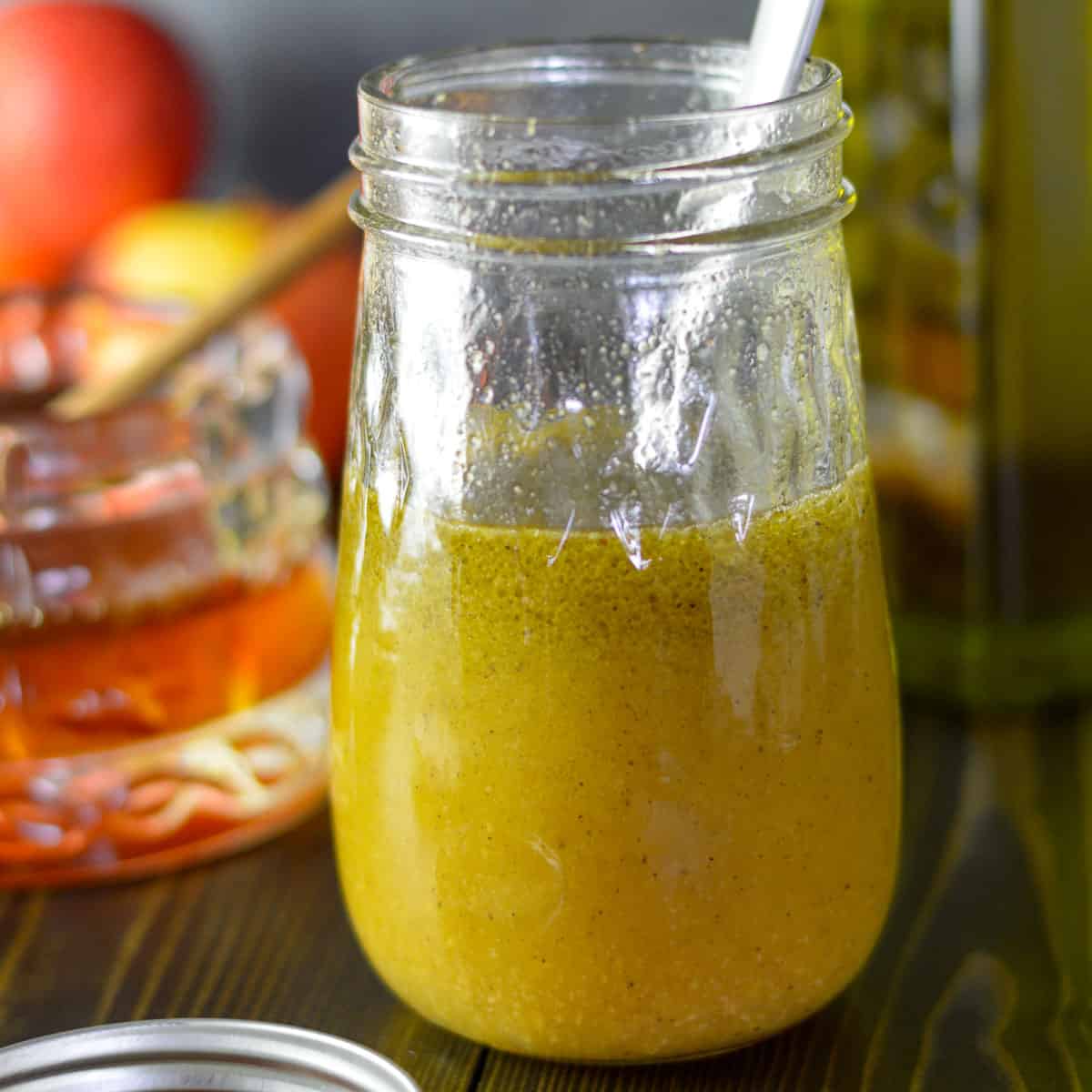 Apple cider vinaigrette salad dressing in a mason jar with a small spoon; honey and olive oil and apples in the background