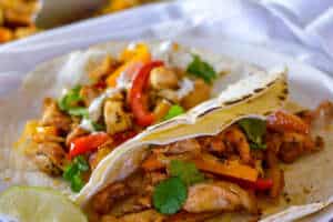 Close up of chicken fajitas in flour tortillas on white plate with a lime wedge and sheet pan of fajitas in background on white napkins