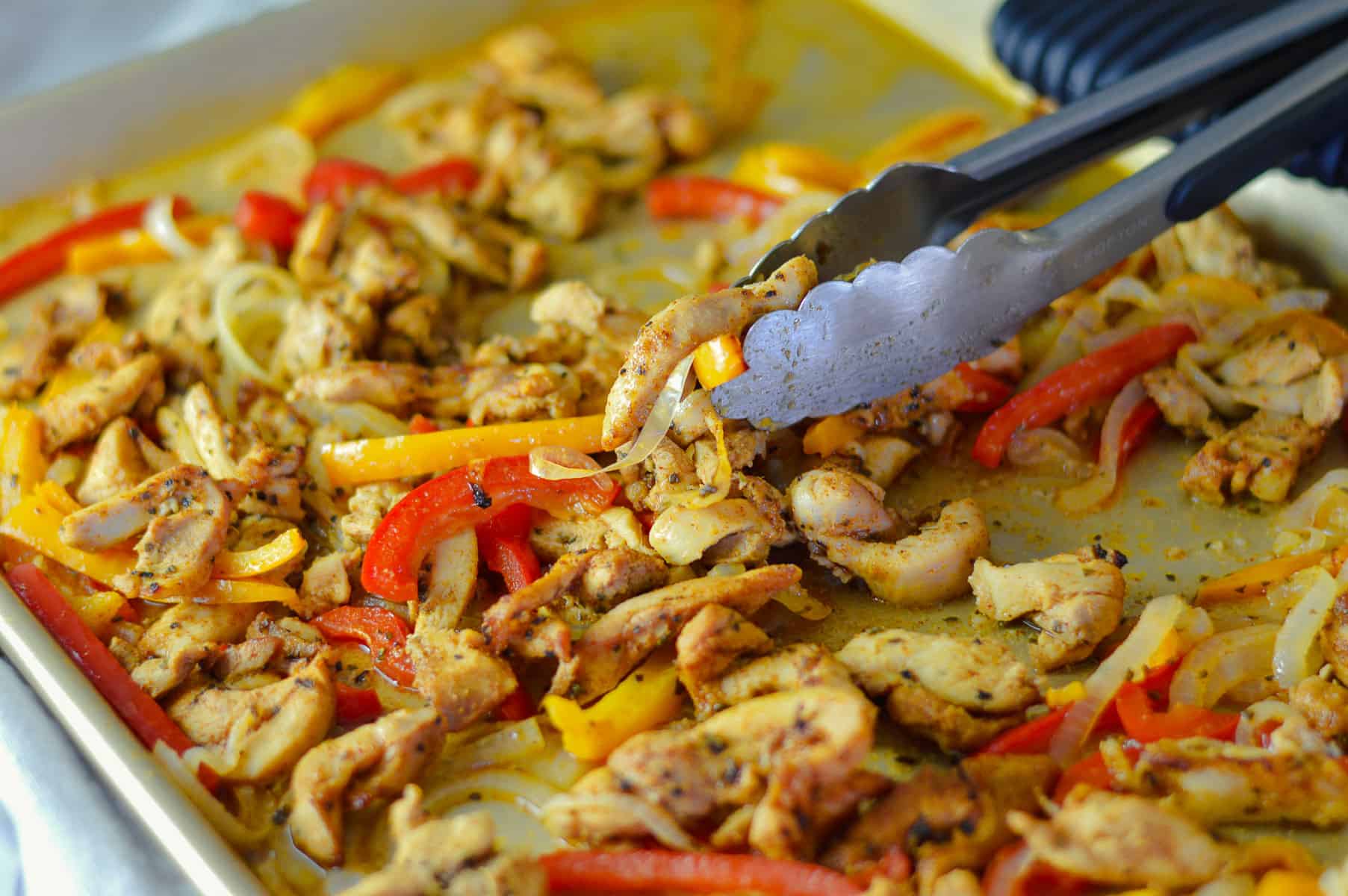 chicken fajita mixture after baking and broiling on gold sheet pan with silver tongs holding some of the mixture
