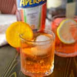 Aperol bourbon cocktail in clear rocks glass with sphere craft ice cubes and candied orange slice with Aperol bottle in background with another cocktail