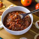 Tomato and Onion Chutney in a white bowl with wooden spoon on top of wooden board with cheese and chutney canapes and tomatoes and onion in the background