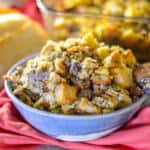 Up close shot of sourdough stuffing with sausage and herbs in a blue and whit bowl with a wooden serving next to a loaf of sourdough bread and a casserole dish of stuffing in the background, all of a red napkin