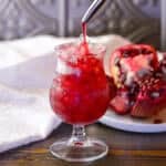 Homemade Grenadine Pomegranate Simple Syrup Featured image showing grenadine being poured into a small fluted glass with crushed ise next to white napkin and an opened pomegranate beside it on a small white plate.