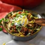 No Bean Chili with Steak and Chorizo in a ceramic bowl with a bronze spoon, sour cream, green onions and shredded cheese with a white napkin, coral napkin behind and red Dutch oven in the back
