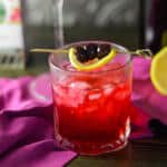 hibiscus mocktail in rocks glass over crushed ice, garnished with a lemon slice and 3 cocktail cherries on wooden table with fuchsia napkin, lemon half and box of tea in background