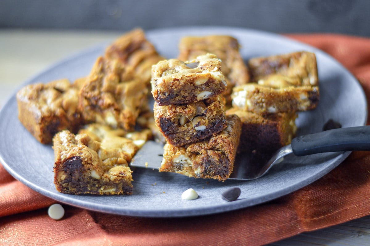 Side shot of blondies on a grey ceramic plate, 3 being held up on a small silver and black handled spatula with an orange shimmery napkin underneath the plate