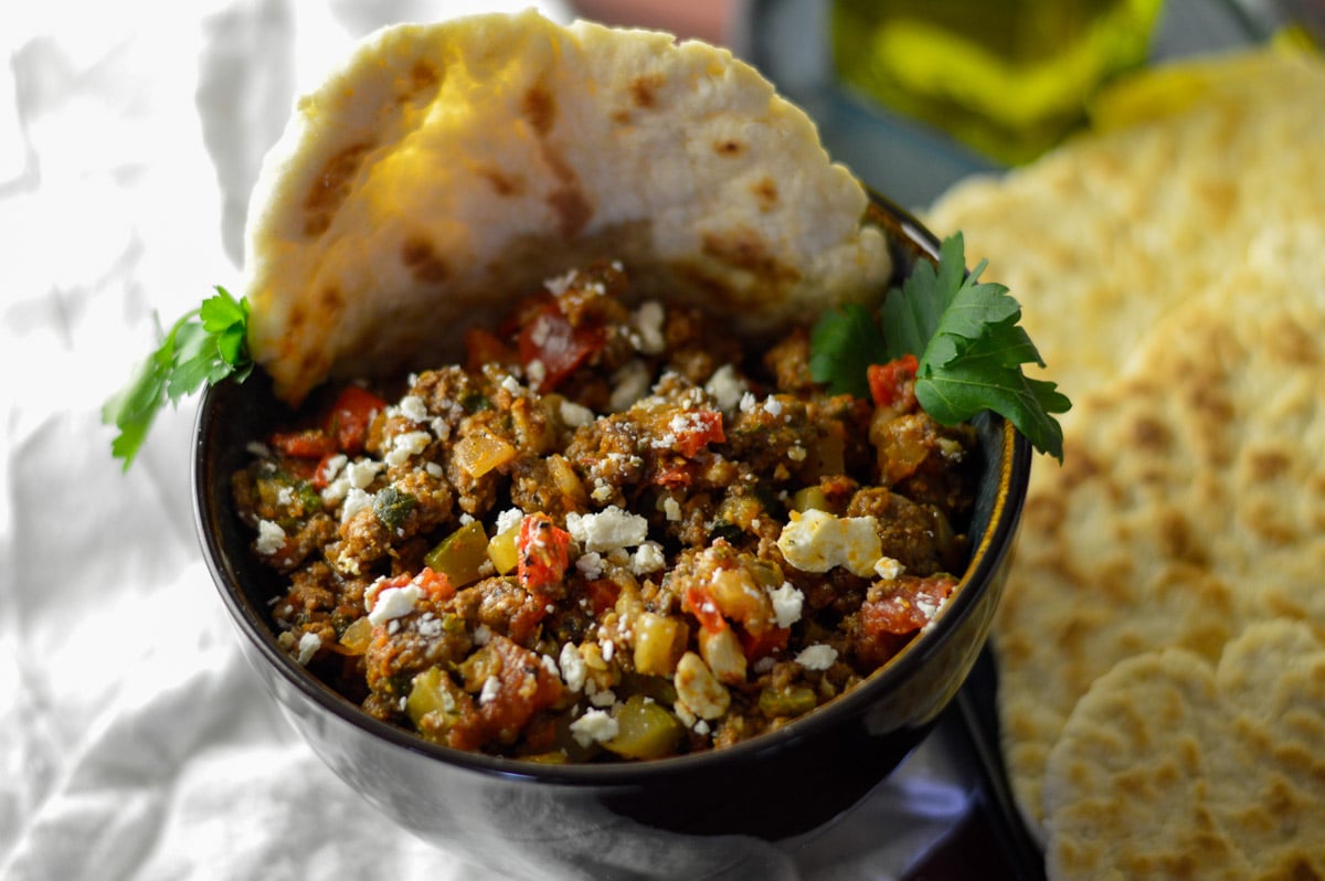 ground beef stir fry dish in a dark ceramic bowl with flatbread, topped with some parsley and feta cheese, more flatbreads on the side.